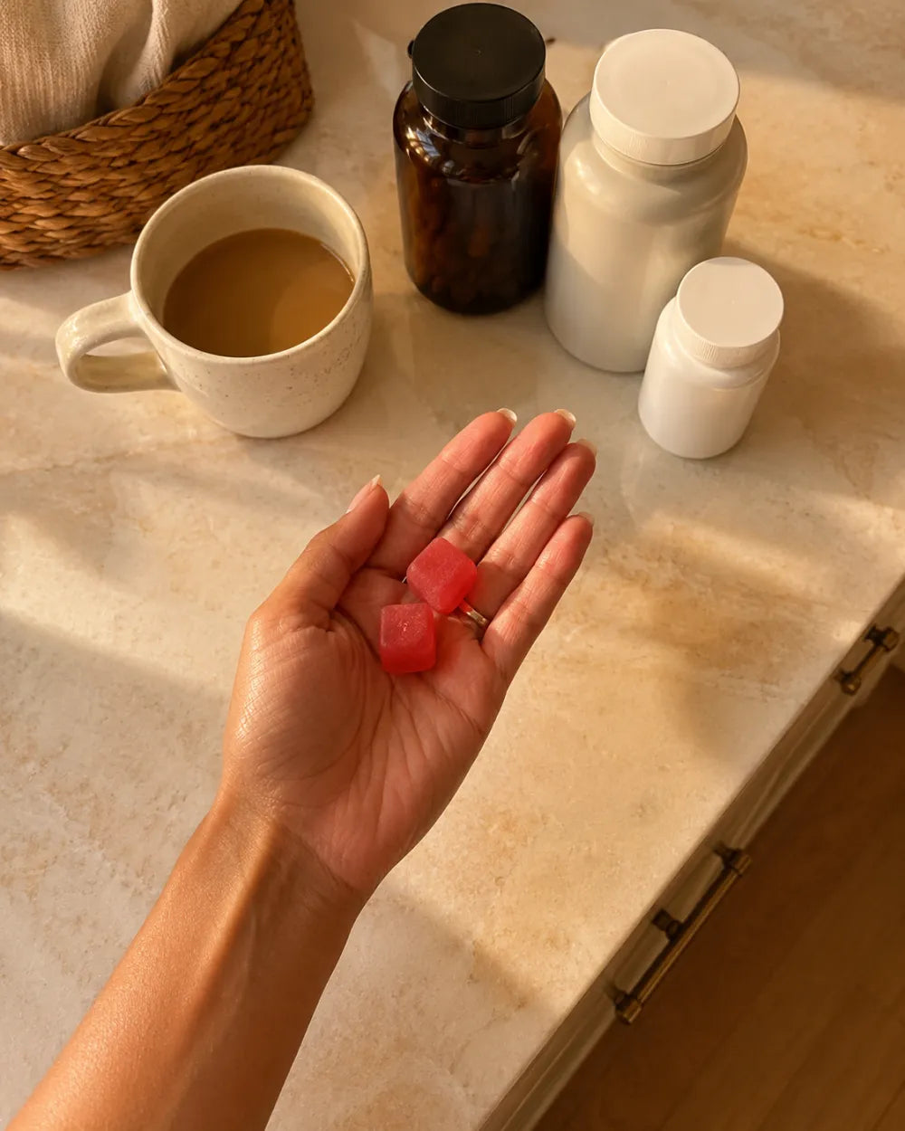 Two gummies in hand beside morning coffee and vitamin bottles